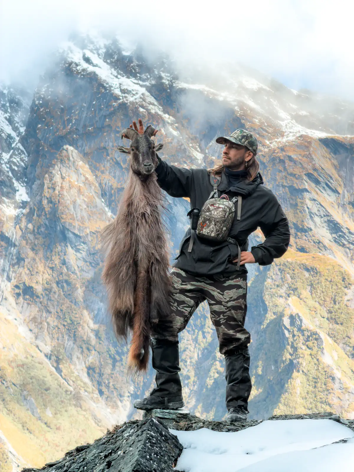 A hunter on a snowy rock ridge holding up a caped bull tahr by the horns, with steep mountains and cloud behind